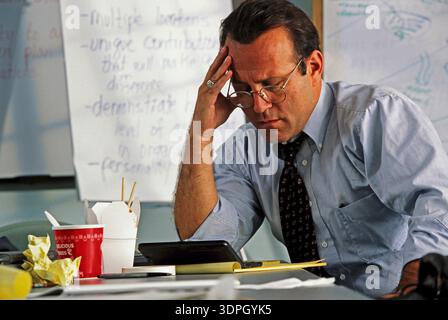 Un uomo si siede alla sua scrivania, mentre lavora, sembra stressato. Tiene la testa in una mano e fissa la sua calcolatrice. Il cibo da asporto si siede accanto a lui Foto Stock
