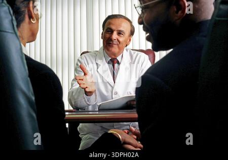 Un medico con un cappotto bianco è seduto ad una scrivania e parla con una giovane coppia. Sembrano essere impegnati in una conversazione sulla salute e il trattamento o Foto Stock