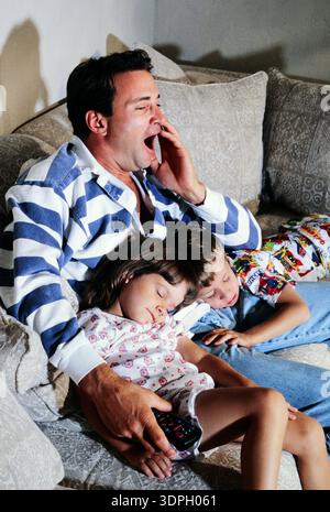 Un padre sbadigliante, assonnato, esausto siede su un divano con i suoi due figli. Un bambino dorme in grembo mentre l'altro riposa accanto a lui. Il padre te Foto Stock
