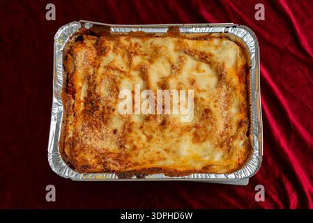 Vista dall'alto verso il basso di lasagne appena sfornate con crosta di formaggio spumante dorata in un vassoio di alluminio monouso, posizionato su un dorso in velluto rosso Foto Stock
