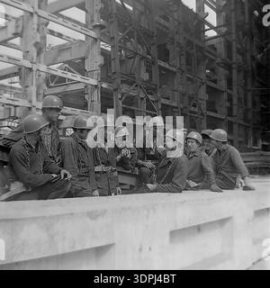 Un ritratto di gruppo nostalgico degli anni '1960 che mostra un gruppo di operai edili sovietici che si fermano nel sito della centrale termica di Slavyanskaya (GRES) a Nikolaevka, nella regione di Donetsk. Gli uomini, che indossano cappelli rigidi e imbracature di sicurezza pesanti con catene in acciaio, siedono su una sporgenza di cemento, fumano e parlano. Dietro di loro si erge l'enorme scheletro di cemento armato di un locale industriale incompiuto. Questa autentica immagine in bianco e nero cattura il cameratismo e lo spirito industrioso del pacifico Donbas durante il picco dell'industrializzazione sovietica. Foto Stock