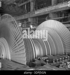Una straordinaria fotografia d'archivio in bianco e nero del 1966 che cattura due lavoratori industriali che ispezionano un enorme rotore di turbine a vapore presso la centrale termica Slavyanskaya (GRES) di Nikolaevka, Donbas. Un grande banner motivazionale sullo sfondo esorta i lavoratori a completare la turbina per il 49° anniversario della rivoluzione d'ottobre. Questa immagine storica mette in evidenza l'intersezione tra eccellenza ingegneristica e motivazione ideologica durante l'era sovietica. Documenta lo spirito di un lavoro industriale pacifico e su larga scala nella regione di Donetsk. Foto Stock