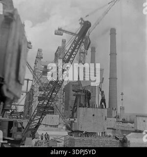 Una straordinaria fotografia d'archivio in bianco e nero del 1965 che cattura un momento cruciale nel cantiere della centrale termica di Slavyanskaya (GRES) a Nikolaevka, Donbas. Una gru cingolata solleva un enorme segnale motivazionale su una base di cemento. Lo striscione delinea obiettivi specifici di lavoro per il collettivo Metallomontazh per quanto riguarda l'unità di potenza da 800 MW, fissando scadenze per ottobre 1965. Questa immagine storica mette in evidenza l'intersezione tra ingegneria e concorrenza socialista del lavoro nello sviluppo industriale pacifico della regione. Foto Stock