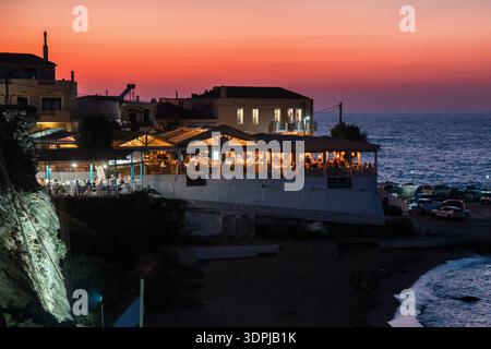 Panormos, Creta, Grecia - 6 settembre 2025: Ristoranti sul mare e porticciolo al tramonto con luci calde che si riflettono sull'acqua nel villaggio costiero Foto Stock
