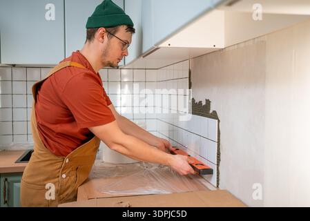 Lavoratore che installa nuove piastrelle da cucina, applica adesivo alla parete, controlla la livella con una livella a bolla d'aria per un progetto di miglioramento della casa perfetto e preciso Foto Stock