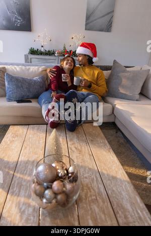 Coppia asiatica seduta sul divano di casa con cappello di Babbo Natale che tiene le tazze accanto alla ciotola con ornamenti in metallo Foto Stock