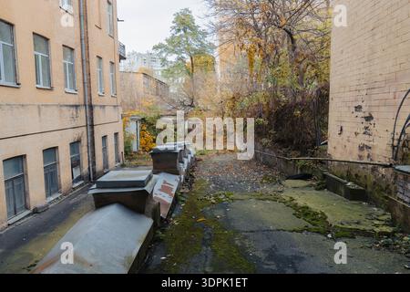 Veduta di uno stretto cortile abbandonato tra vecchi edifici gialli in autunno. Pavimentazione ricoperta di muschio verde e foglie cadute in un vicolo cittadino Foto Stock