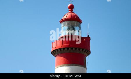 Un faro bianco con una distintiva cupola rossa si erge alto contro un cielo azzurro limpido, mostrando la classica architettura marittima. Foto Stock