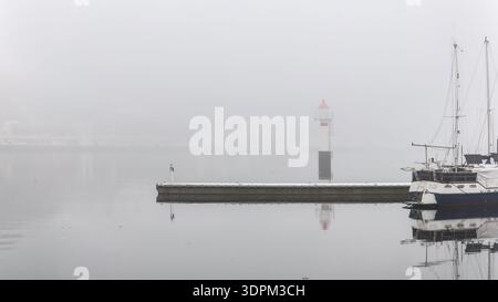 Silenziosa quiete, minimalismo nordico, barca a vela ormeggiata accanto al molo galleggiante con un piccolo faro in una fitta nebbia costiera sull'acqua calma per la costa locale Foto Stock