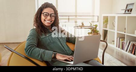 La giovane donna lavora su un computer portatile in una camera moderna durante il giorno mentre è seduta sulla sedia Foto Stock