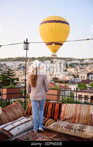 Una donna si erge sulla terrazza panoramica dell'hotel con cuscini a motivi geometrici e tappeti intrecciati, guardando la mongolfiera volare sopra la città all'alba Foto Stock