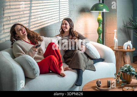 Due allegre donne amiche sedute su un divano, ridendo e sorridendo mentre passavano il tempo libero insieme in un luminoso soggiorno Foto Stock
