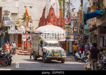 Udaipur, India - 23 marzo 2022: Un camion decorato percorre una strada stretta con pedoni, motociclette, scooter e negozi commerciali, passando di fronte a un tempio indù con uno shikhara rosso e bianco durante il giorno. Foto Stock