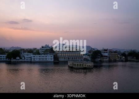 Udaipur, India - 23 marzo 2022: Gli edifici storici sul lungomare e il padiglione Gangaur Ghat sono visibili sul bordo del lago Pichola al crepuscolo, con montagne sullo sfondo e un cielo pastello sopra la testa. Foto Stock