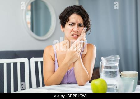 Giovane donna con mal di denti e guancia toccante, seduta a tavola Foto Stock