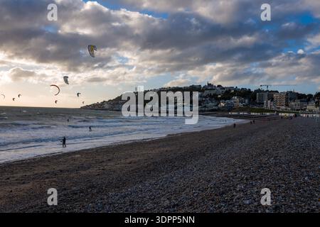 Le Havre, Francia - 25 agosto 2020: Diversi kitesurfer cavalcano le onde sul mare vicino a un'ampia spiaggia di ciottoli, con persone che camminano lungo il litorale, gli edifici della città e le case in collina visibili sullo sfondo sotto un cielo nuvoloso. Foto Stock