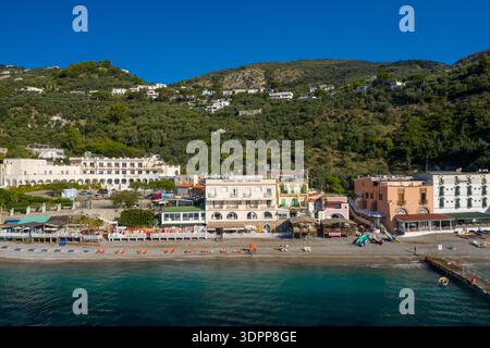 Marina del Cantone, Italia - 6 ottobre 2020: L'immagine mostra una fila di edifici e alberghi sul lungomare lungo la spiaggia di ciottoli di Marina del Cantone, con colline coperte di verde che si innalzano sullo sfondo sotto un cielo azzurro. Foto Stock