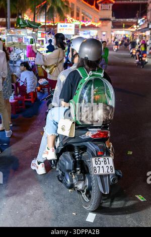 CAN Tho, Vietnam - 19 gennaio 2020: Una donna che indossa un casco guida uno scooter con un piccolo cane all'interno di uno zaino trasparente, navigando attraverso un affollato mercato notturno con bancarelle illuminate, venditori e acquirenti visibili sullo sfondo. Foto Stock