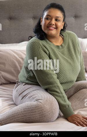 Donna asiatica di mezza età seduta a gambe incrociate sul letto con maglione verde in maglia e pantaloni a costine Foto Stock