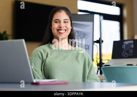 Donna ispanica sorridente seduta su un tavolo dai colori chiari in uno studio di design con laptop e planimetrie Foto Stock