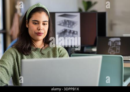 Donna che lavora su un notebook argentato, due monitor che mostrano modelli 3D, con sedia verde in studio di design Foto Stock