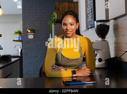 Barista afroamericana che si piega e piega le braccia al bancone in un bar con tablet e smerigliatrice Foto Stock
