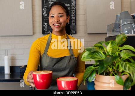 Barista afro-americano sorridente che indossa la mostarda e il grembiule verde che serve tazze di latte dietro il bar Foto Stock