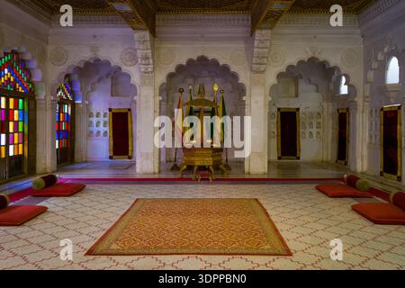 Jodhpur, India - 25 marzo 2022: L'immagine mostra una sala di ricevimento al forte di Mehrangarh caratterizzata da un trono centrale con bandiere, vetrate colorate, porte ad arco, cuscini rossi e tappeti a motivi geometrici. Foto Stock