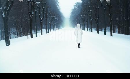 Donna con un piumino bianco che cammina su un sentiero innevato attraverso un buio vicolo della foresta invernale. Il freddo nebbioso crea un'atmosfera misteriosa Foto Stock