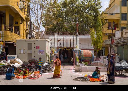 Udaipur, India - 23 marzo 2022: Diverse donne si siedono e stanno in piedi per strada vendendo fiori e offerte a terra vicino all'ingresso di un tempio indù, con statue di elefanti decorate, motociclette ed edifici residenziali visibili sullo sfondo. Foto Stock
