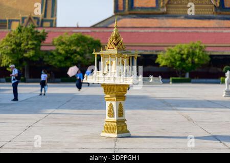 Phnom Penh, Cambogia - 6 febbraio 2020: Un santuario in miniatura ornato sorge su un piedistallo decorato nel cortile pavimentato aperto del complesso del Palazzo reale, con edifici in stile Khmer, arbusti curati e diverse persone visibili sullo sfondo. Foto Stock