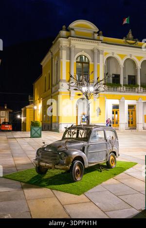 Ville de Verallo, Italia - 27 settembre 2020: Una vecchia scultura di auto con rami di alberi metallici che emergono dal suo tetto è esposta su erba artificiale in una piazza lastricata di notte, con un edificio giallo e bianco e bandiera italiana sullo sfondo. Foto Stock
