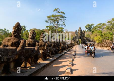 Entree de Angkor Thom, Cambogia - 14 febbraio 2020: Statue di guardiani di pietra fiancheggiano entrambi i lati del ponte che conduce al cancello d'ingresso di Angkor Thom, con persone che cavalcano tuk e motociclette sulla strada sotto la luce del giorno. Foto Stock