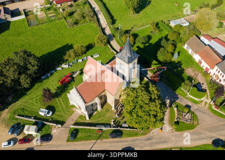 Cuncy, Francia - 30 settembre 2021: Una vista aerea mostra una chiesa in pietra con un tetto piastrellato rosso e un campanile, circondato da auto parcheggiate, persone vicino all'ingresso, attrezzature da costruzione e case del villaggio in un paesaggio rurale. Foto Stock