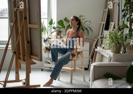Ritrattista donna concentrata che dipinge su cavalletto in uno studio domestico creativo pieno di piante. Foto Stock