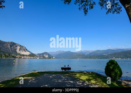Isola dei pescatori, Italia - 30 settembre 2020: Due persone sono sedute su una panchina di legno ai margini di una zona lacustre erbosa e ghiaiosa, affacciata sul Lago maggiore con montagne lontane e un cielo azzurro limpido visibile. Foto Stock