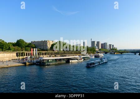 Parigi, Francia - 10 maggio 2022: Diversi battelli fluviali sono attraccati lungo la Senna a Port de Suffren con persone che camminano sul lungofiume, moderni edifici alti e uno storico edificio in pietra visibile sullo sfondo sotto la luce del sole. Foto Stock