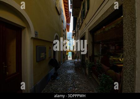 Isola dei pescatori, Italia - 30 settembre 2020: Uno stretto vicolo acciottolato fiancheggiato da edifici residenziali gialli e color crema presenta una vetrina con interni visibili, persiane verdi, piante in vaso e una vista sul Lago maggiore in lontananza. Foto Stock