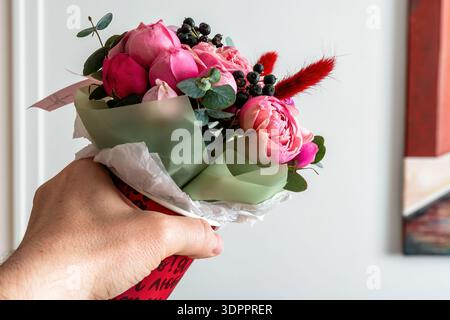 Mano dell'uomo che regge un bellissimo bouquet rosa con frutti di bosco ed eucalipto in una tazza rossa decorativa, perfetta per un regalo romantico o una celebrazione Foto Stock
