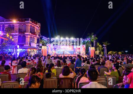 Chau Doc, Vietnam - 24 gennaio 2020: Una grande folla è seduta su sedie di plastica e guarda un palco all'aperto illuminato con decorazioni festose e cartellonistica durante un evento notturno del capodanno cinese, con edifici a più piani e palme visibili sullo sfondo. Foto Stock