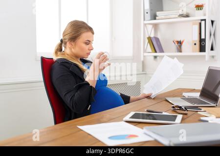 Donna incinta che legge documenti e beve caffè alla sua scrivania durante le ore diurne Foto Stock