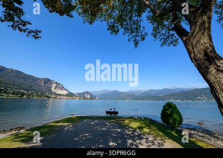 Isola dei pescatori, Italia - 30 settembre 2020: Due persone siedono su una panchina di legno ai margini di un'area lacustre erbosa e ghiaiosa, affacciata sul Lago maggiore con montagne lontane e un cielo azzurro limpido visibile. Foto Stock