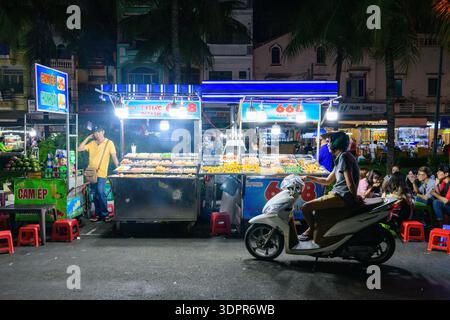 CAN Tho, Vietnam - 19 gennaio 2020: I venditori di cibo gestiscono bancarelle illuminate che vendono vari spuntini e bevande di notte, con un uomo in moto in primo piano e gruppi di persone sedute su sgabelli rossi nelle vicinanze. Foto Stock