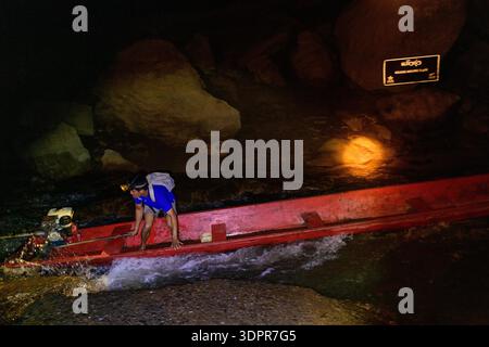 Grotte de Konglor, Laos - 3 giugno 2020: Un uomo con una camicia blu e un faro, manovra una barca a coda lunga rossa con un motore attraverso acque poco profonde e rocce all'interno di una grotta scura, con un cartello di avvertimento illuminato visibile sullo sfondo. Foto Stock