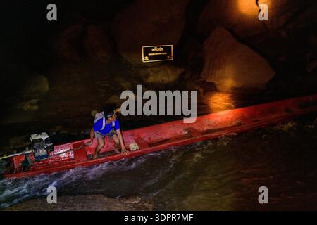 Grotte de Konglor, Laos - 3 giugno 2020: Un uomo che indossa una camicia blu e un faro manovra una barca a coda lunga rossa con un motore attraverso acque poco profonde e rocce all'interno di una grotta scura, con un cartello di avvertimento illuminato visibile sullo sfondo. Foto Stock
