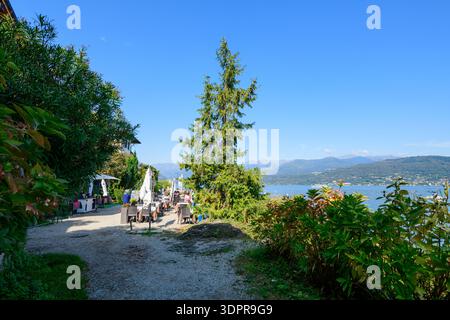Isola dei pescatori, Italia - 30 settembre 2020: Tavoli all'aperto con persone sedute sotto gli ombrelloni sono disposti lungo un sentiero ghiaioso lungo la riva del lago, con cespugli, un albero di conifere e vista sul Lago maggiore e sulle montagne lontane sotto un cielo azzurro. Foto Stock