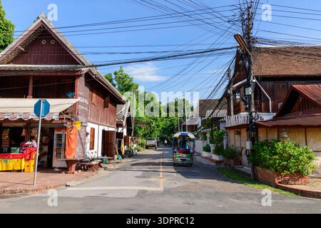 Luang Prabang, Laos - 30 giugno 2020: Tradizionali edifici in legno a due piani, un piccolo negozio sulla strada, un tuk-tuk e cavi elettrici sospesi visibili sono visti lungo una strada tranquilla con vegetazione sotto un cielo azzurro. Foto Stock