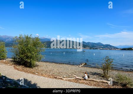 Isola dei pescatori, Italia - 30 settembre 2020: Una persona siede su un pezzo di mare su una sponda rocciosa e acciottolata con cespugli sparsi, affacciata sul Lago maggiore con montagne lontane e un cielo azzurro. Foto Stock