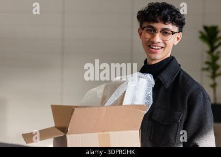 Uomo che indossa una giacca che disimballa la scatola con la bolla in ufficio con la pianta in vaso, spazio per copiare Foto Stock