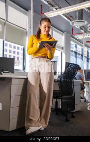 Donna asiatica adulta che indossa la parte superiore della senape appoggiata al piedistallo della scrivania, utilizzando un tablet in ufficio Foto Stock
